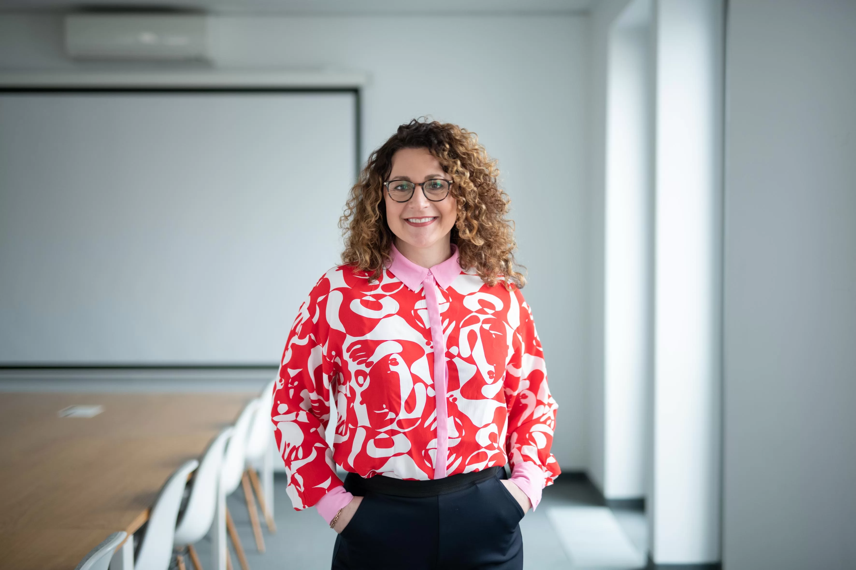 Eine Frau mit Locken und einer bunten Bluse steht in einem Konferenzraum und lächelt. 