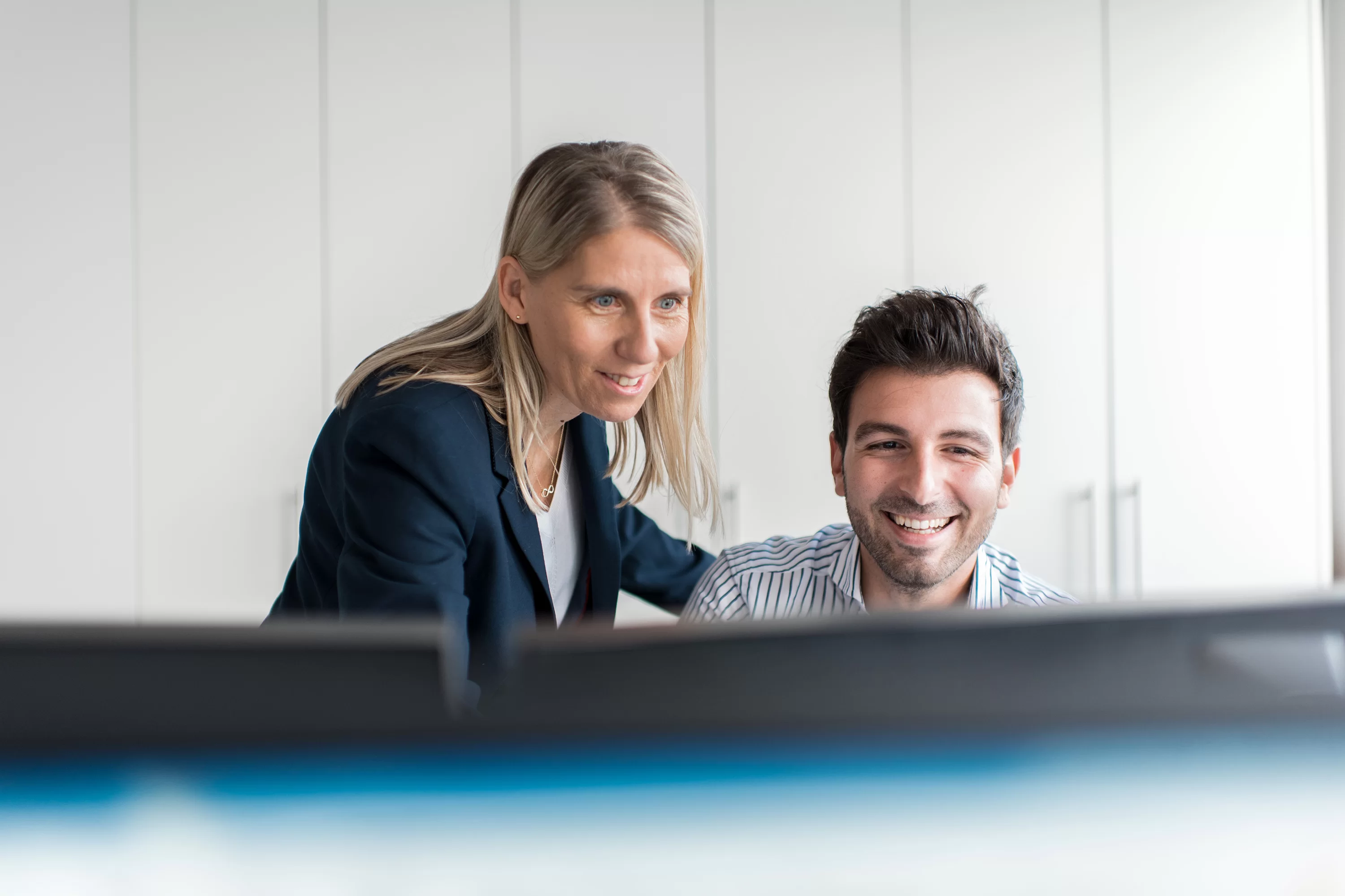 Eine Frau steht am Arbeitsplatz eines Mannes, beide schauen freundlich auf den Monitor