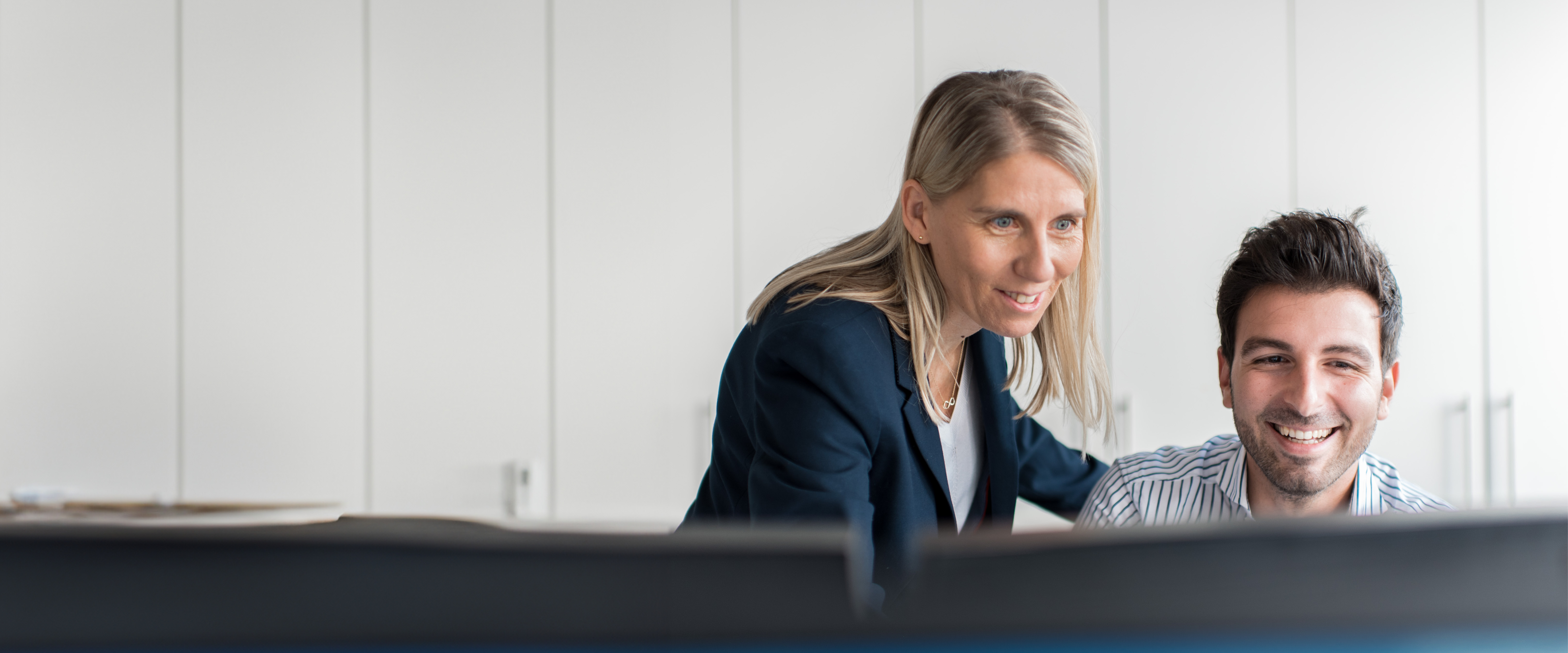 Eine Frau steht am Arbeitsplatz eines Mannes, beide schauen freundlich auf den Monitor