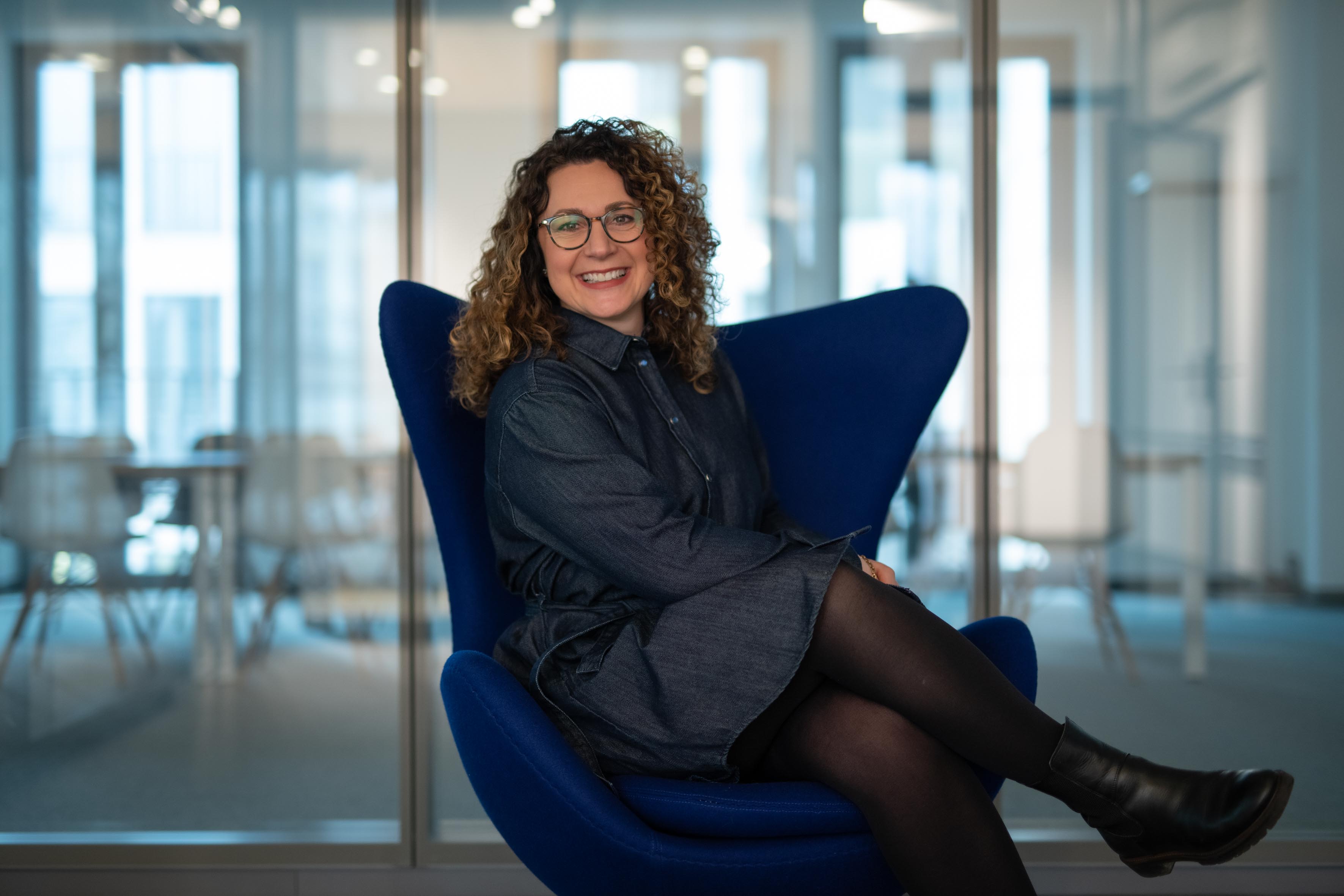 Eine Frau mit Locken und dunkelblauen Kleid sitzt auf einem Stuhl und lacht.