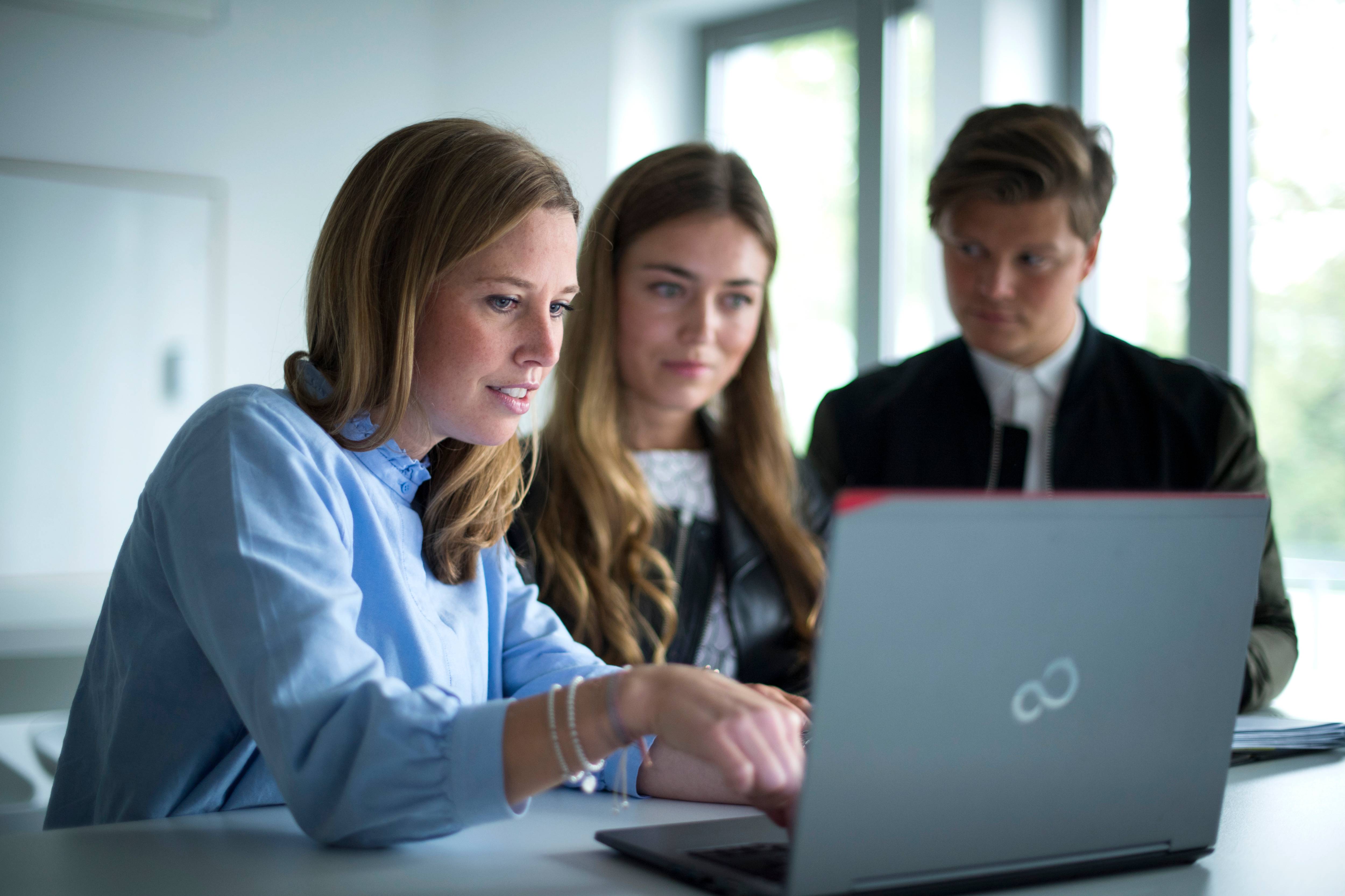 Eine Frau sitzt an einem Laptop und zeigt einer anderen Frau und einem Mann etwas darauf.