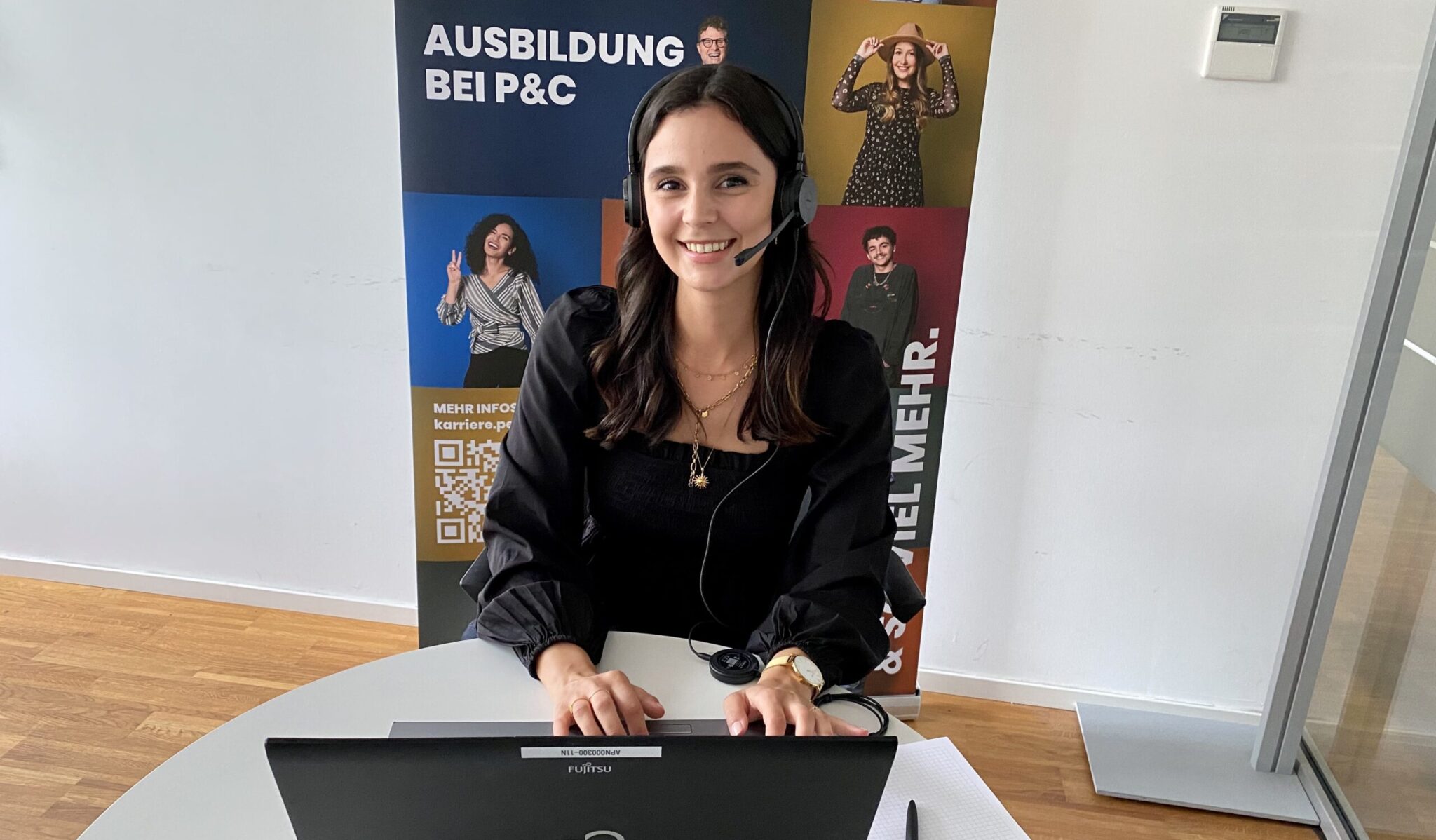 Chiara sitzt mit Headset und Laptop am Schreibtisch, im Hintergrund steht ein bunter P&C Aufsteller.