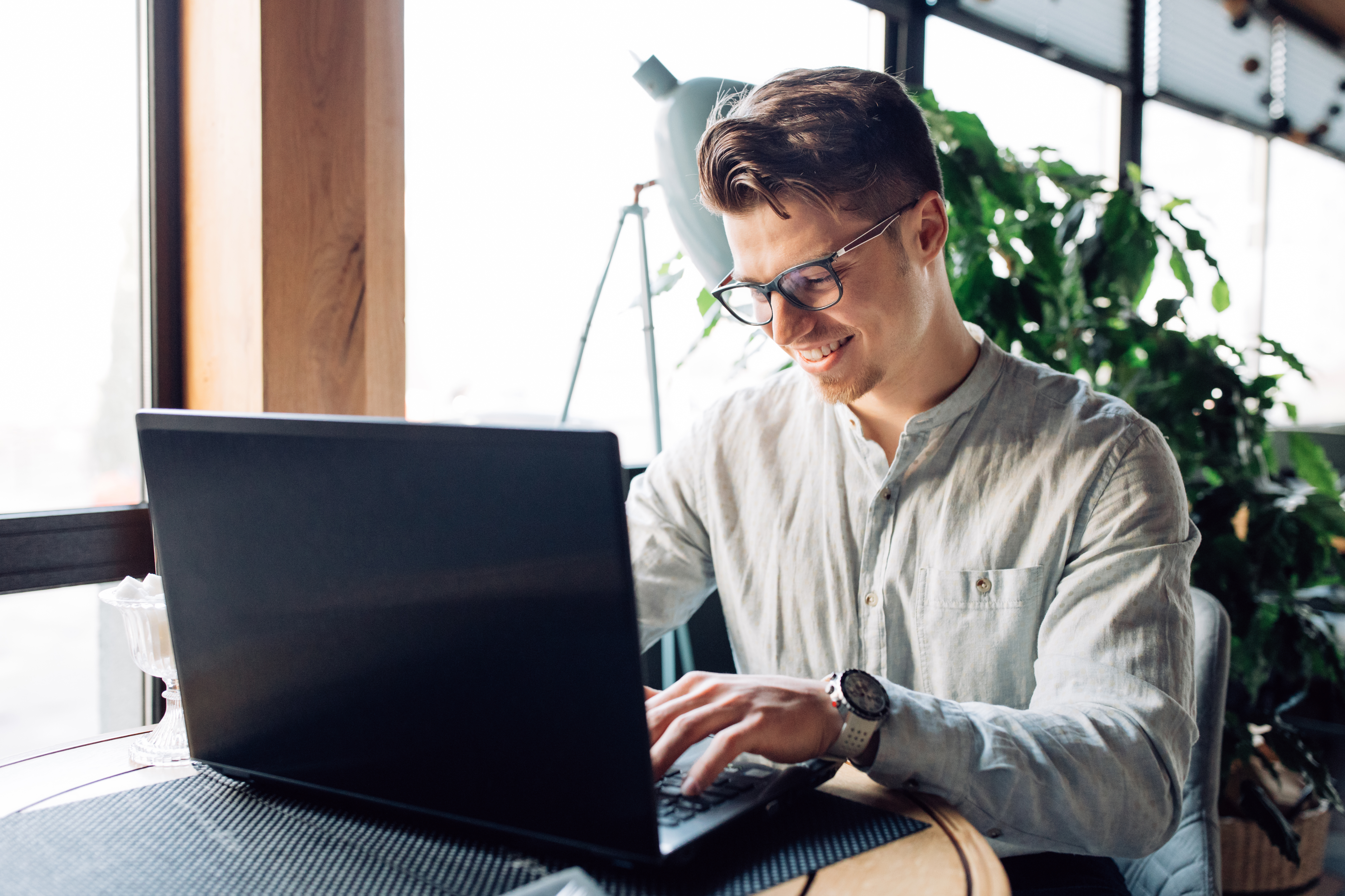 Ein junger Mann mit Brille und Hemd sitzt am Tisch an seinem Laptop und lächelt, während er tippt.