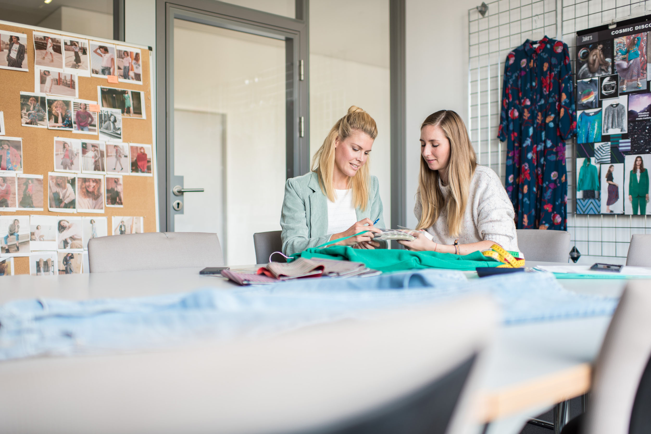 Zwei Frauen sitzen an einem Tisch voller Stoffe und sprechen über die Farbmuster, die sie in der Hand halten.